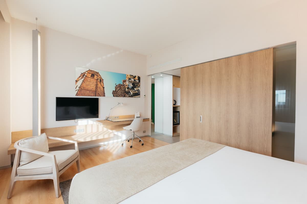 Moderni hotellihuone, jossa on suuri sänky, työpöytä tuolilla, taulutelevisio ja taulu seinällä.