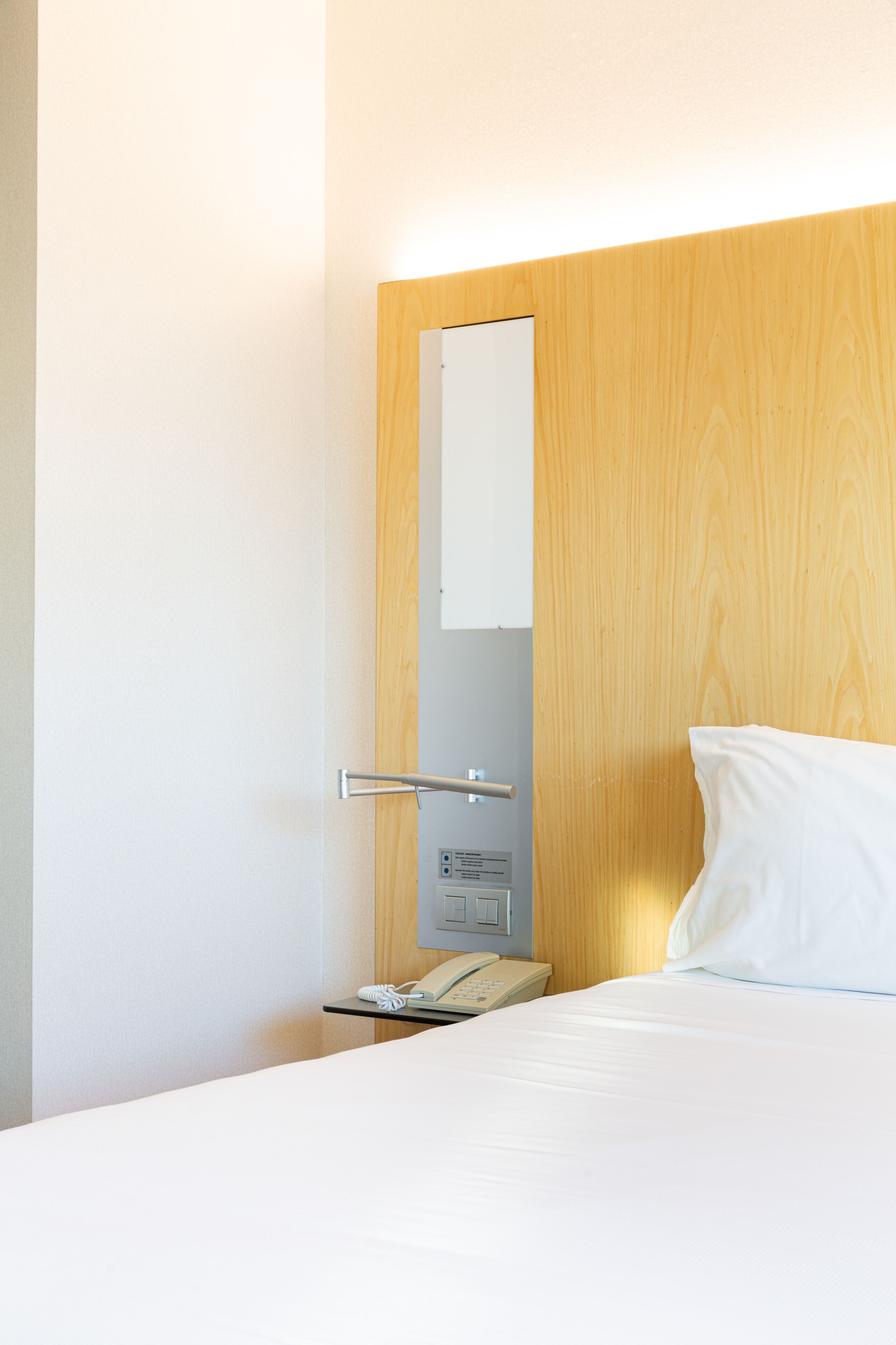 A hotel room with a wooden headboard, a white bed, a bedside table with a telephone, and a wall-mounted light.