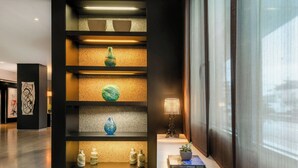 A modern interior with a black shelving unit displaying decorative items, a table with a lamp and a book, and a large window with a view. - AC Hotel Padova by Marriott (Padova)