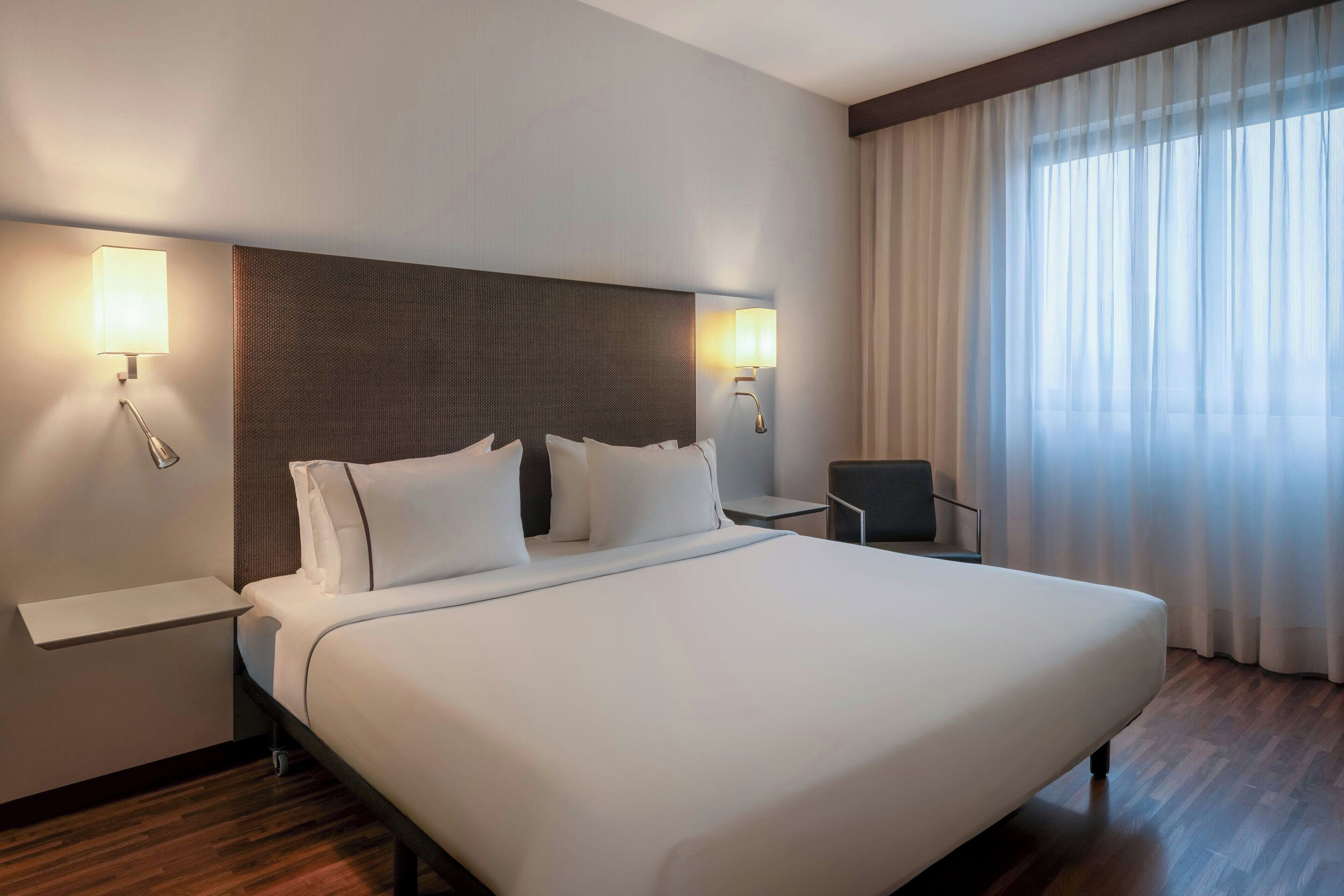 A hotel room with a large bed, two bedside tables, a chair, and a window with curtains.