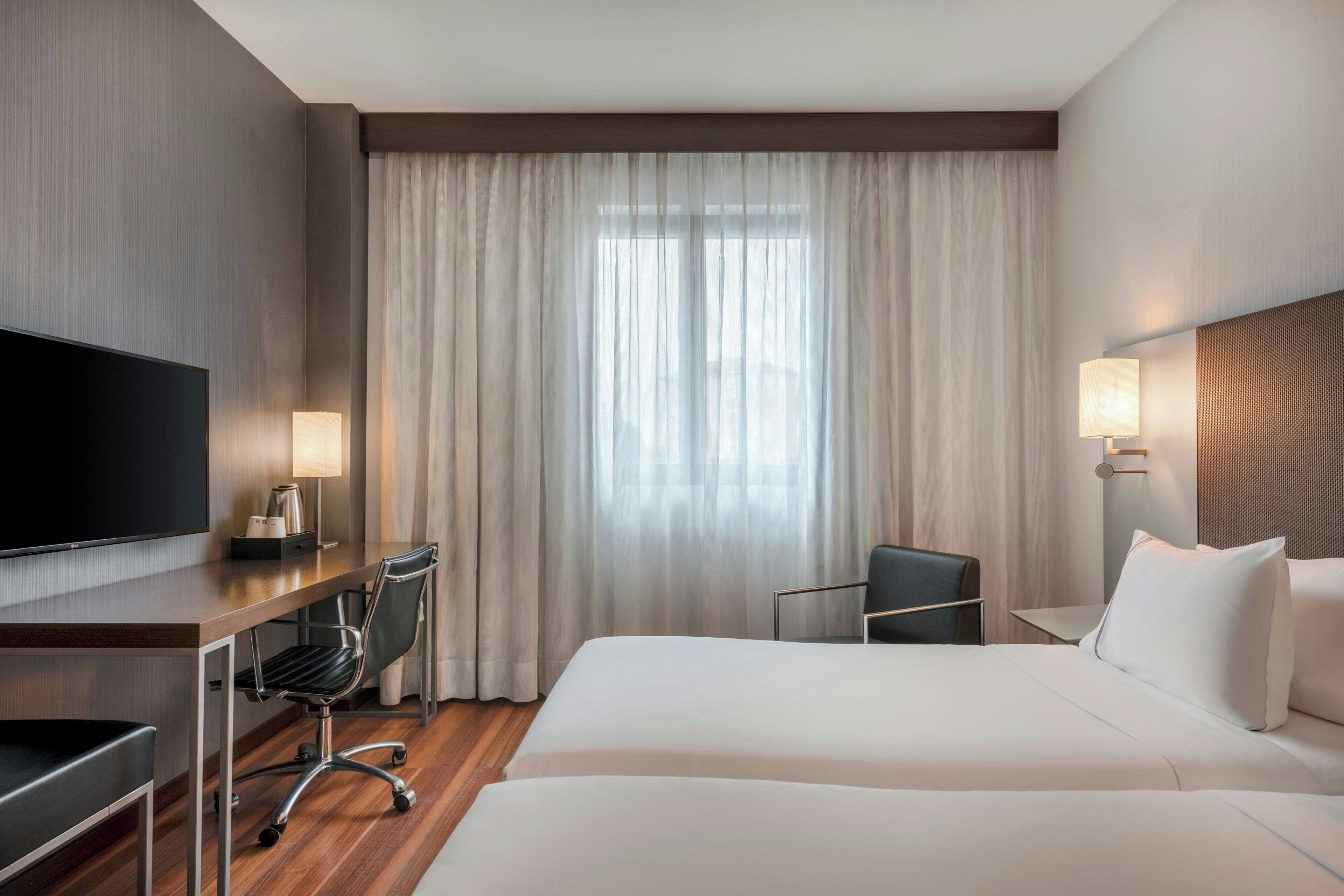 Habitación de hotel con cama, escritorio, silla, televisor y ventana con cortinas.
