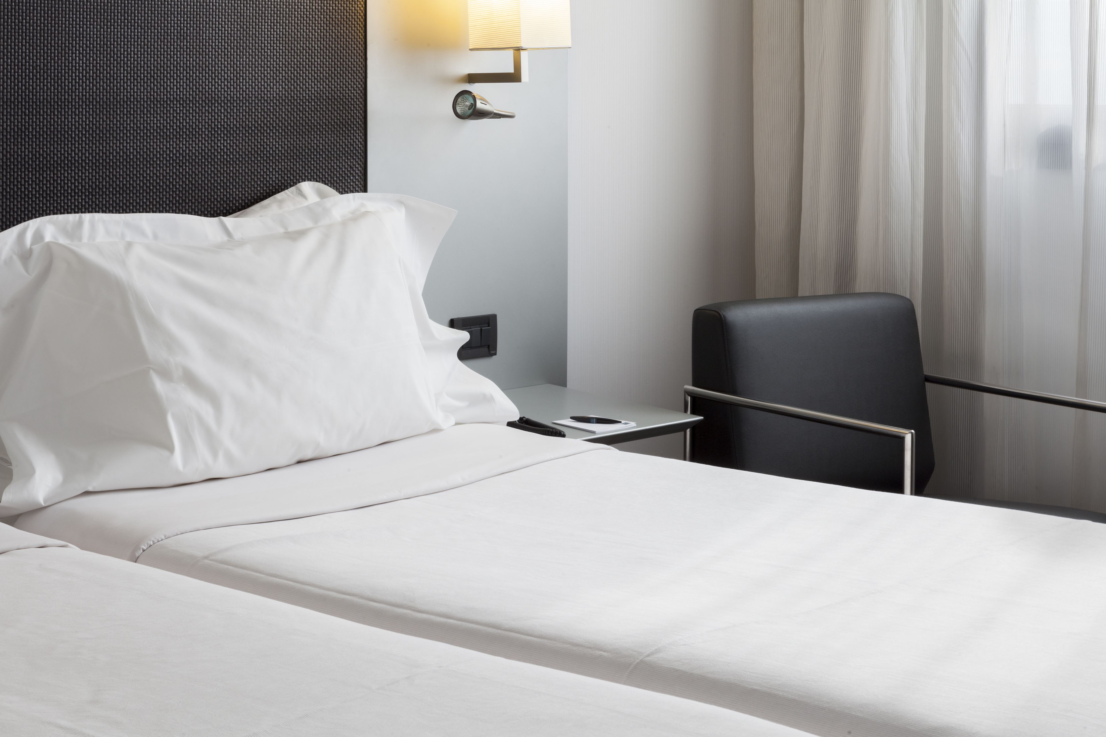A hotel room with a neatly made bed, a small bedside table with a lamp, a chair, and a view of the outside through sheer curtains.