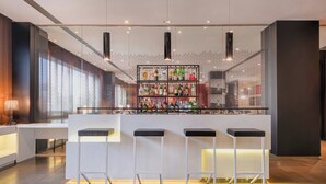 A bar area with a white counter, three stools, and a shelf of various bottles. - AC Hotel Padova by Marriott (Padova)