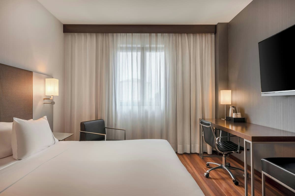 A hotel room with a large bed, a desk, a chair, a TV, and a window with curtains.