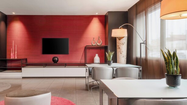 A modern hotel room with a flat-screen TV, a dining area with a table and chairs, a red accent wall, and a large window. - AC Hotel Padova by Marriott (Padova)