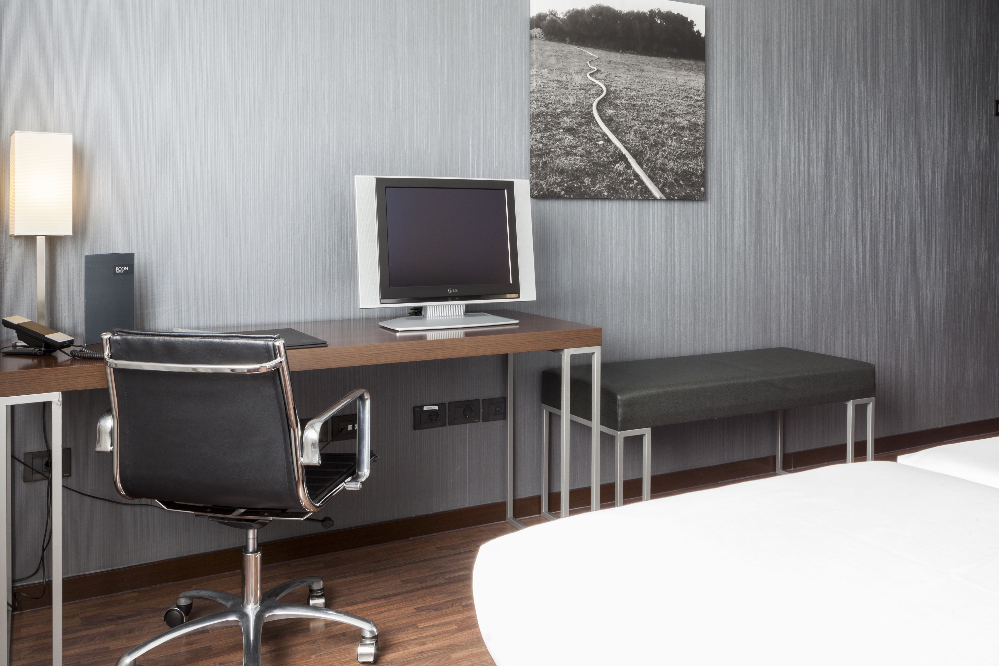 Una habitación de hotel moderna con un escritorio de madera, una silla de oficina negra, un televisor de pantalla plana, un banco y un cuadro en la pared.