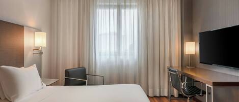 A hotel room with a large bed, a desk with a chair, a TV, and a window with curtains.
