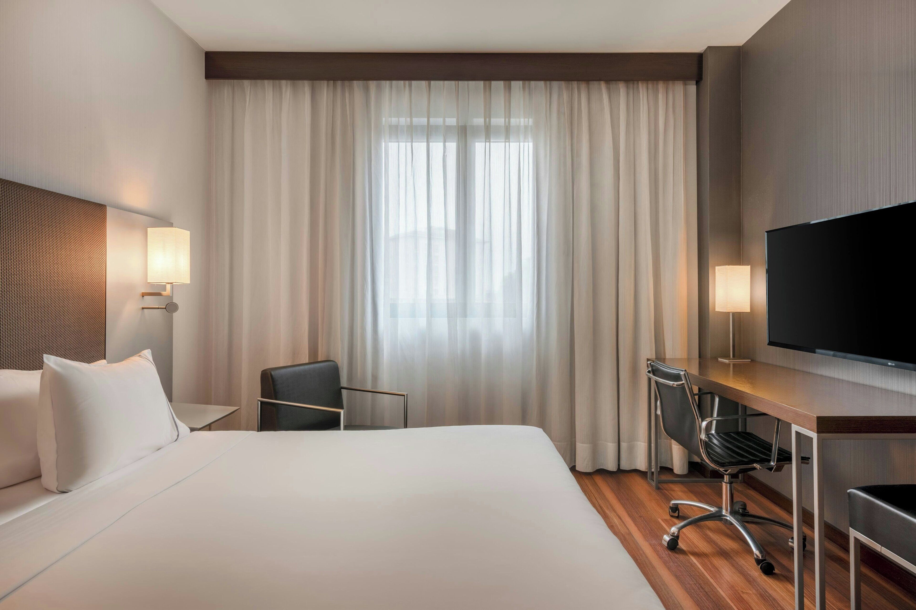 A hotel room with a large bed, a desk with a chair, a television, and a window with curtains.