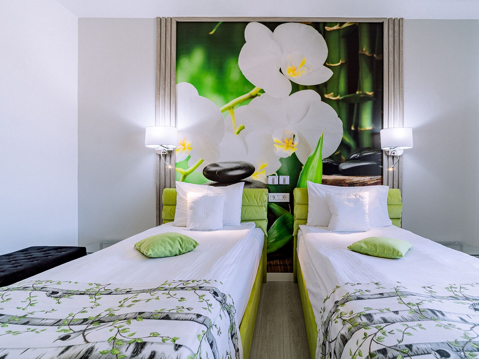 Une chambre d’hôtel avec deux lits, chacun agrémenté d’un oreiller vert et d’une couvre-lit blanche ornée de motifs représentant des branches d’arbres. Des lampes murales sont fixées de chaque côté du lit.