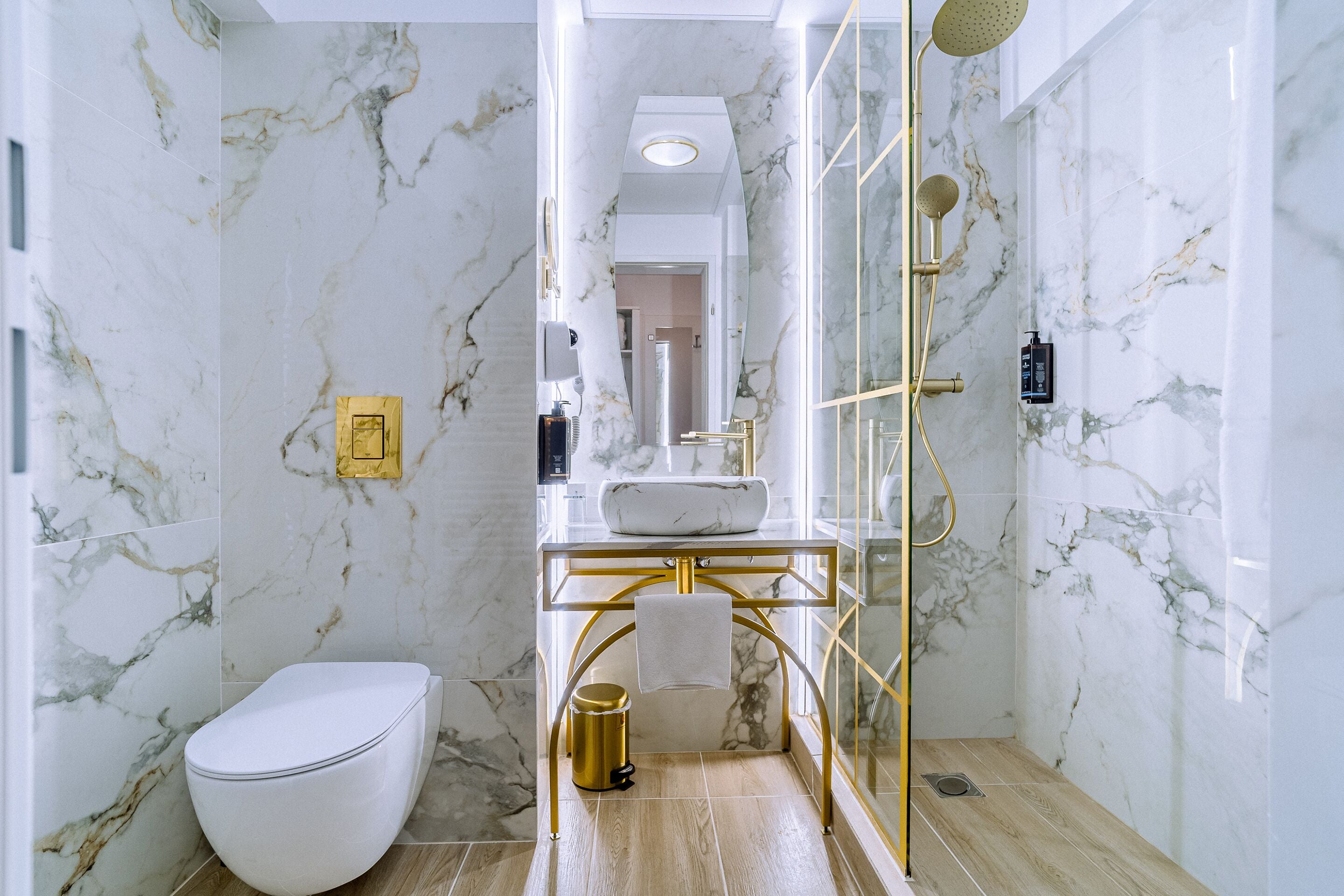 Un baño con pared de mármol, tocador dorado, inodoro blanco y zona de ducha.