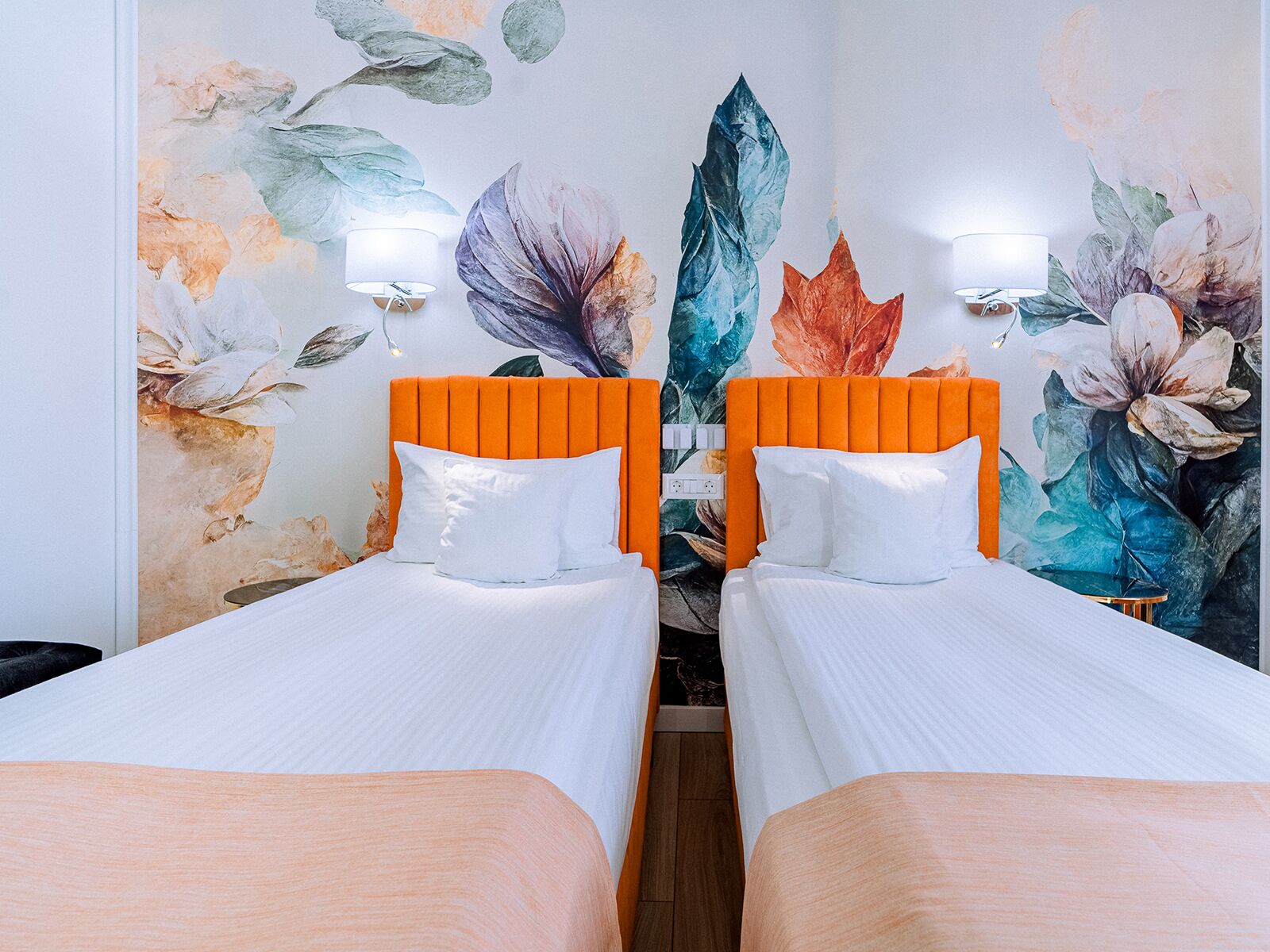 A hotel room with two beds, each with an orange headboard, white bedding, and a floral mural on the wall.