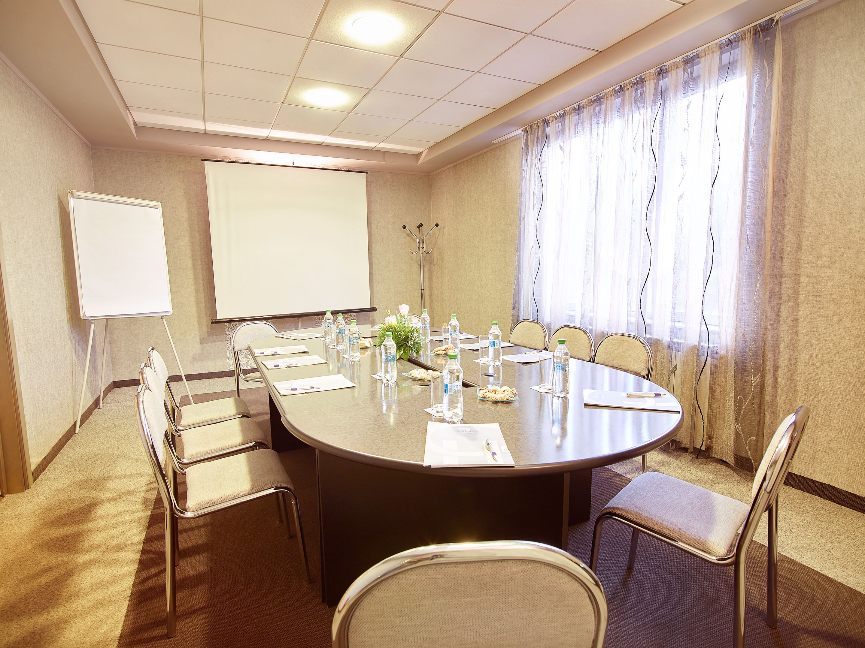 A conference room with a large oval table, chairs, a whiteboard, and a projector screen.
