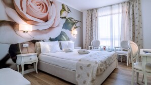 A hotel room with a large floral mural, a bed with white linens, two bedside tables with lamps, a desk with a chair, and a large window with curtains. - Continental Forum Oradea (Oradea)
