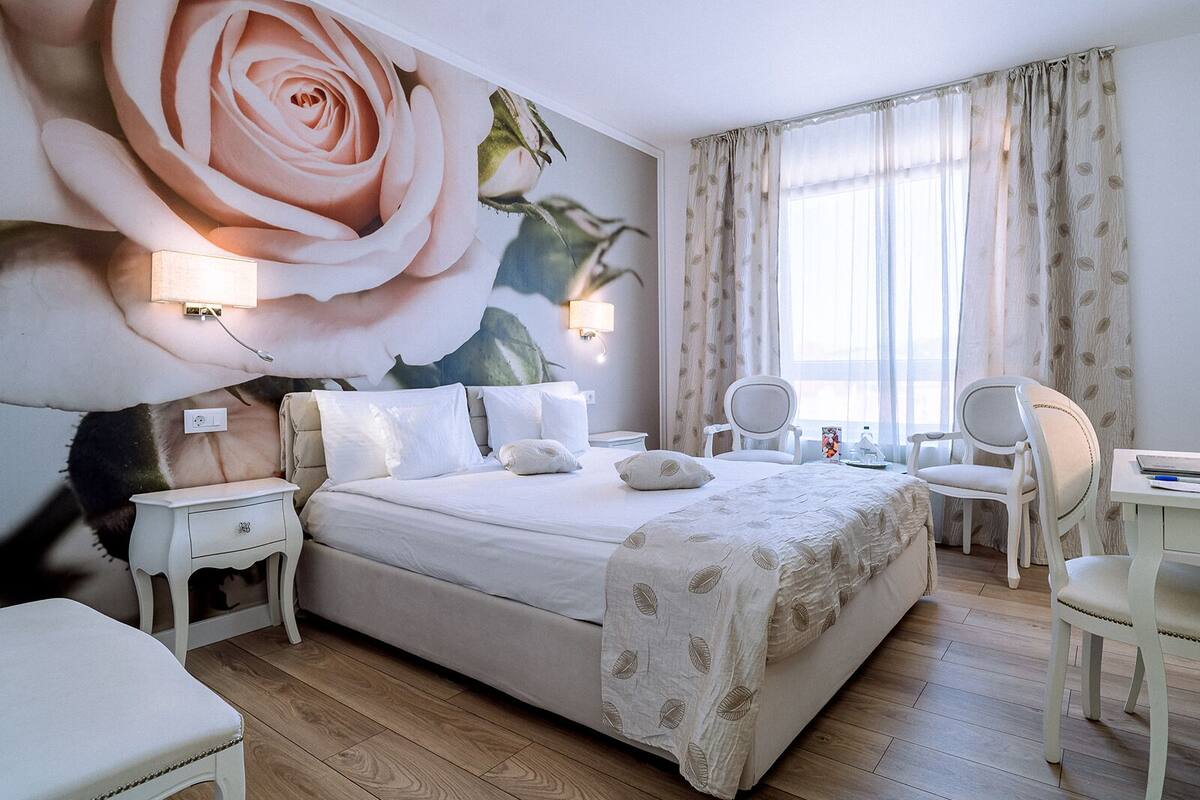 A hotel room with a large floral mural, a bed with white linens, two bedside tables with lamps, a desk with a chair, and a large window with curtains.