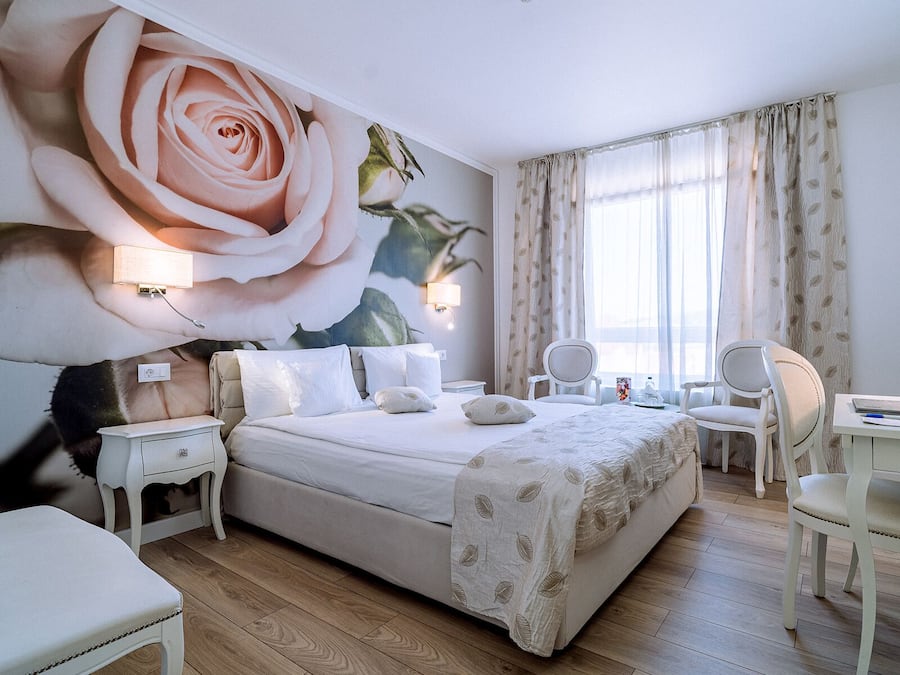 Habitación de hotel con un amplio mural floral, cama con ropa blanca, dos mesitas de noche con lámparas, un escritorio con silla y un ventanal con cortinas.