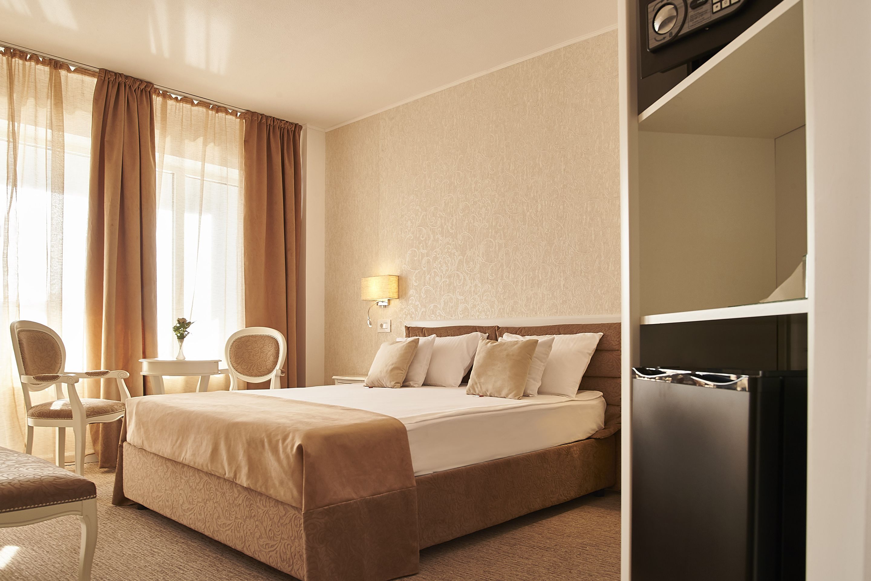 A hotel room with a bed, two chairs, a small table, a lamp, and a built-in refrigerator.