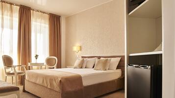 Habitación de hotel con cama, dos sillas, una mesita, lámpara y refrigerador incorporado.