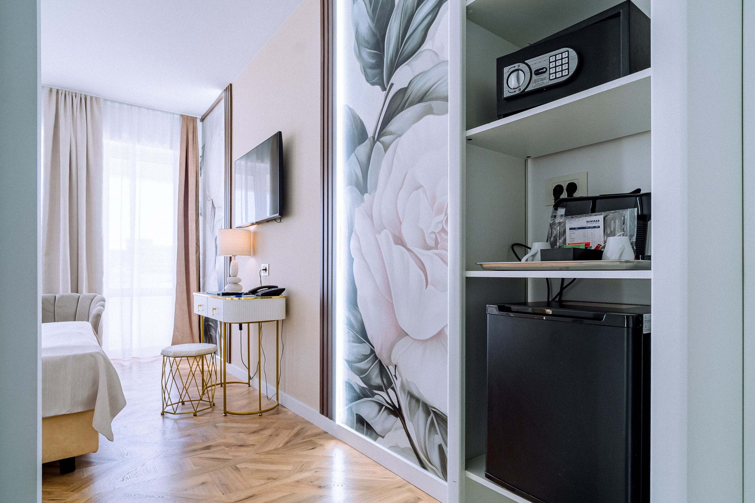 A hotel room with a bed, a desk with a lamp, a refrigerator, and a wall with a floral mural.