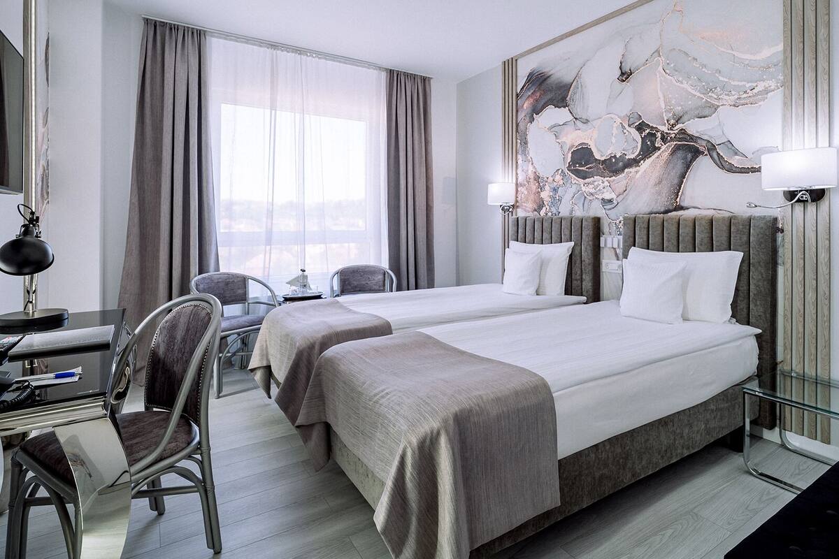 A hotel room with two beds, a desk with a chair, a large window with curtains, and a wall-mounted lamp.