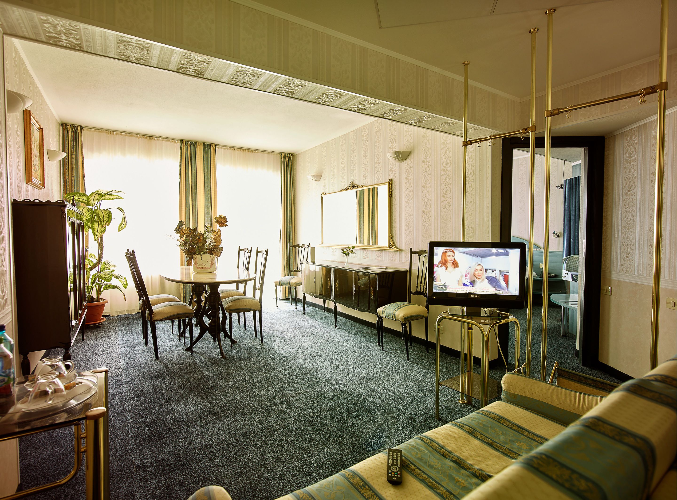 A hotel room with a dining table, chairs, a sofa, a television, and a potted plant.