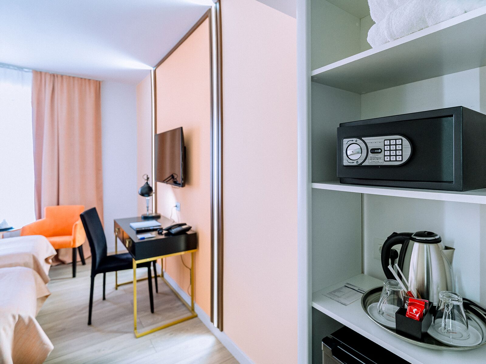 A hotel room with a desk, chair, bed, and a shelf with a coffee maker and a kettle.