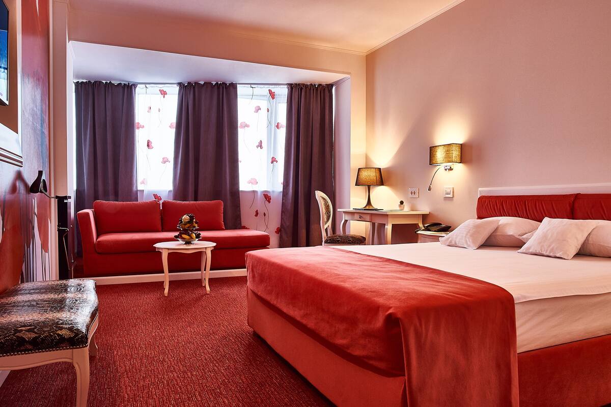 A hotel room with a large bed, a red sofa, a small table, and a chair.
