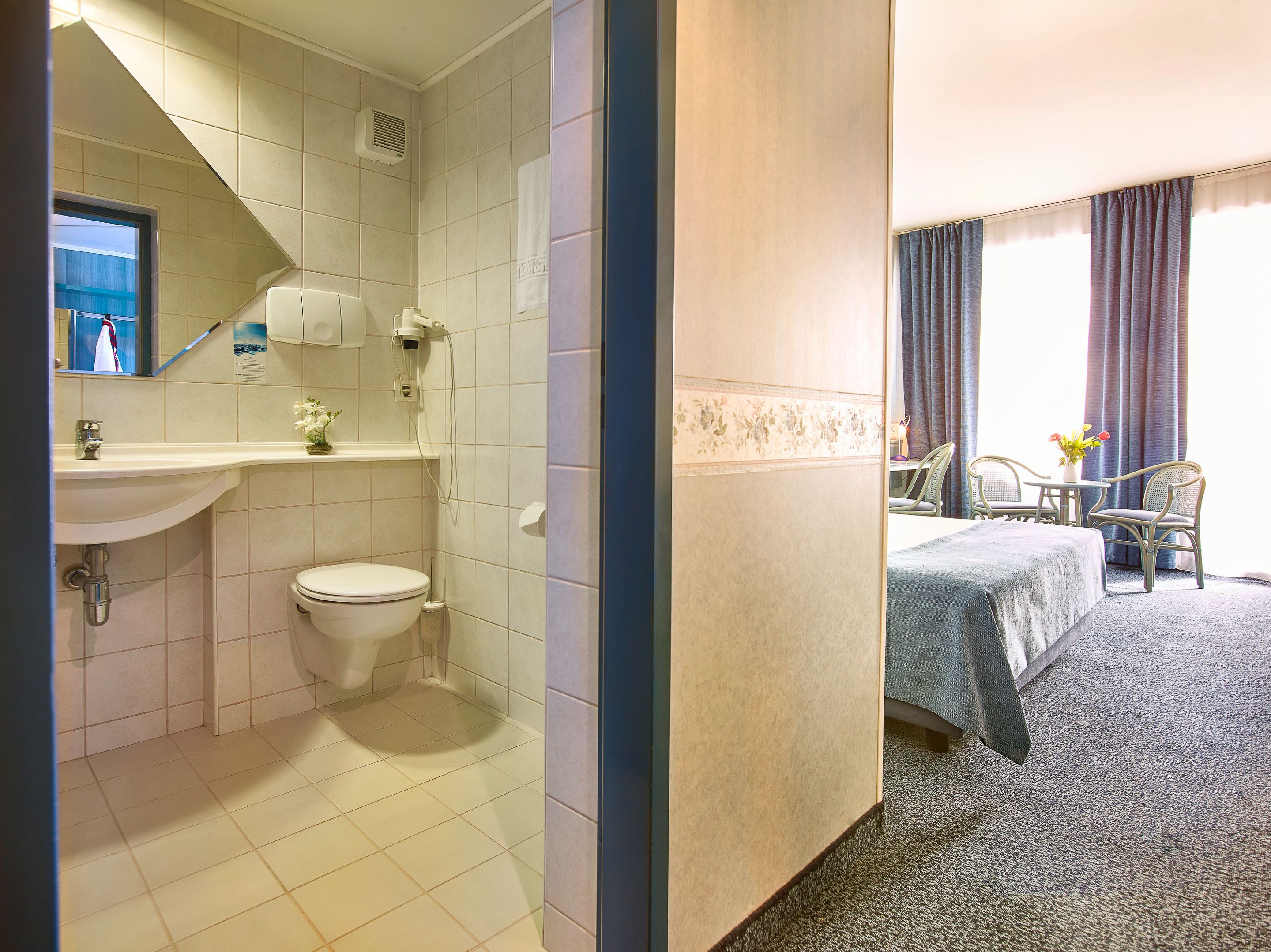 A hotel bathroom with a toilet, sink, and shower. There is a bed with a table and chairs in the adjacent room.