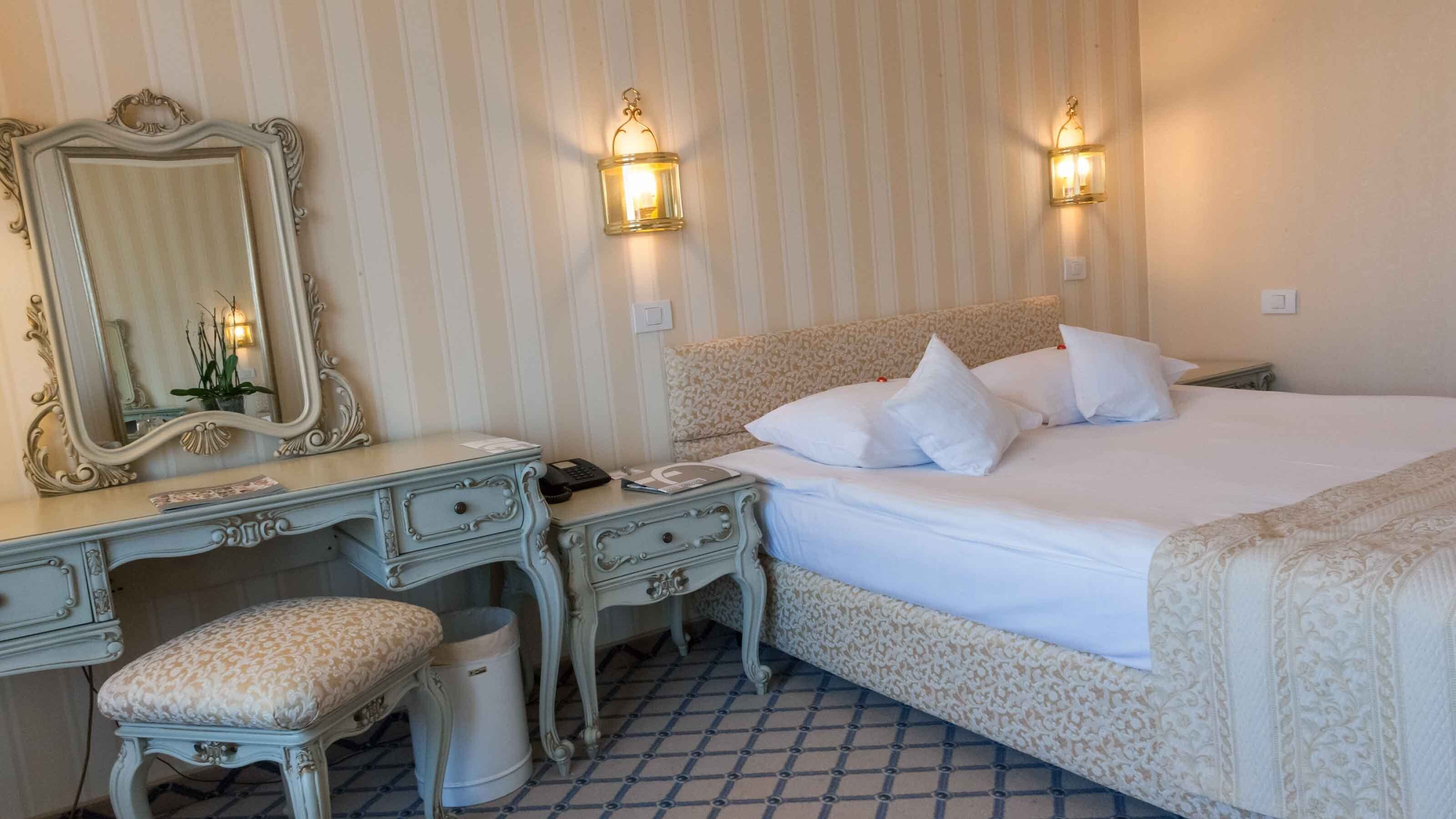 A hotel room with a large bed, a vanity with a mirror, two wall sconces, a small stool, and a trash can.