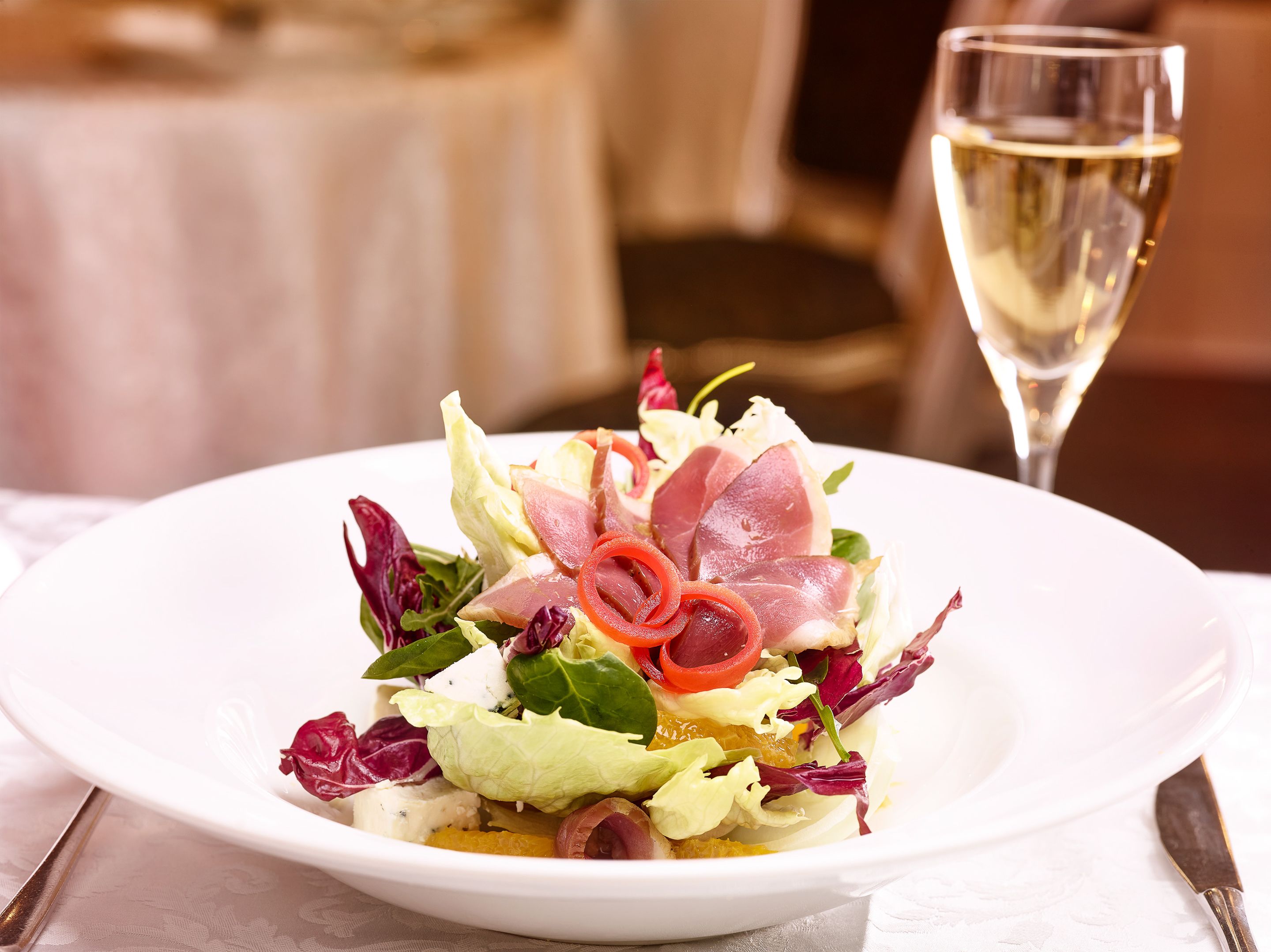 A white plate with a salad, a glass of white wine, and a knife on a table.
