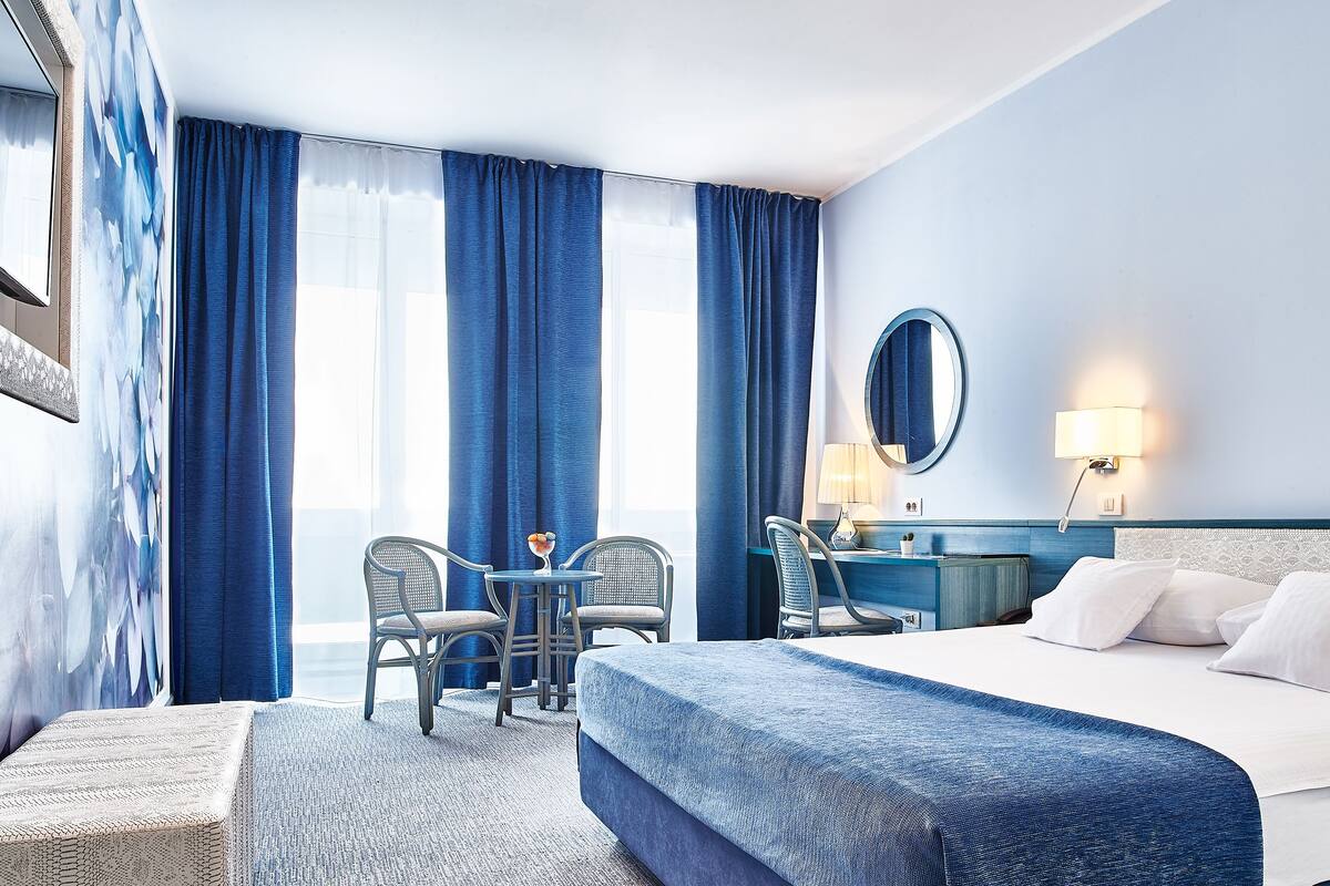 A hotel room with a large bed, a desk with a chair, a small table with a chair, and a large window with curtains.