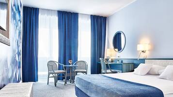 Habitación de hotel con una cama grande, un escritorio con silla, una mesita con otra silla y un ventanal con cortinas.