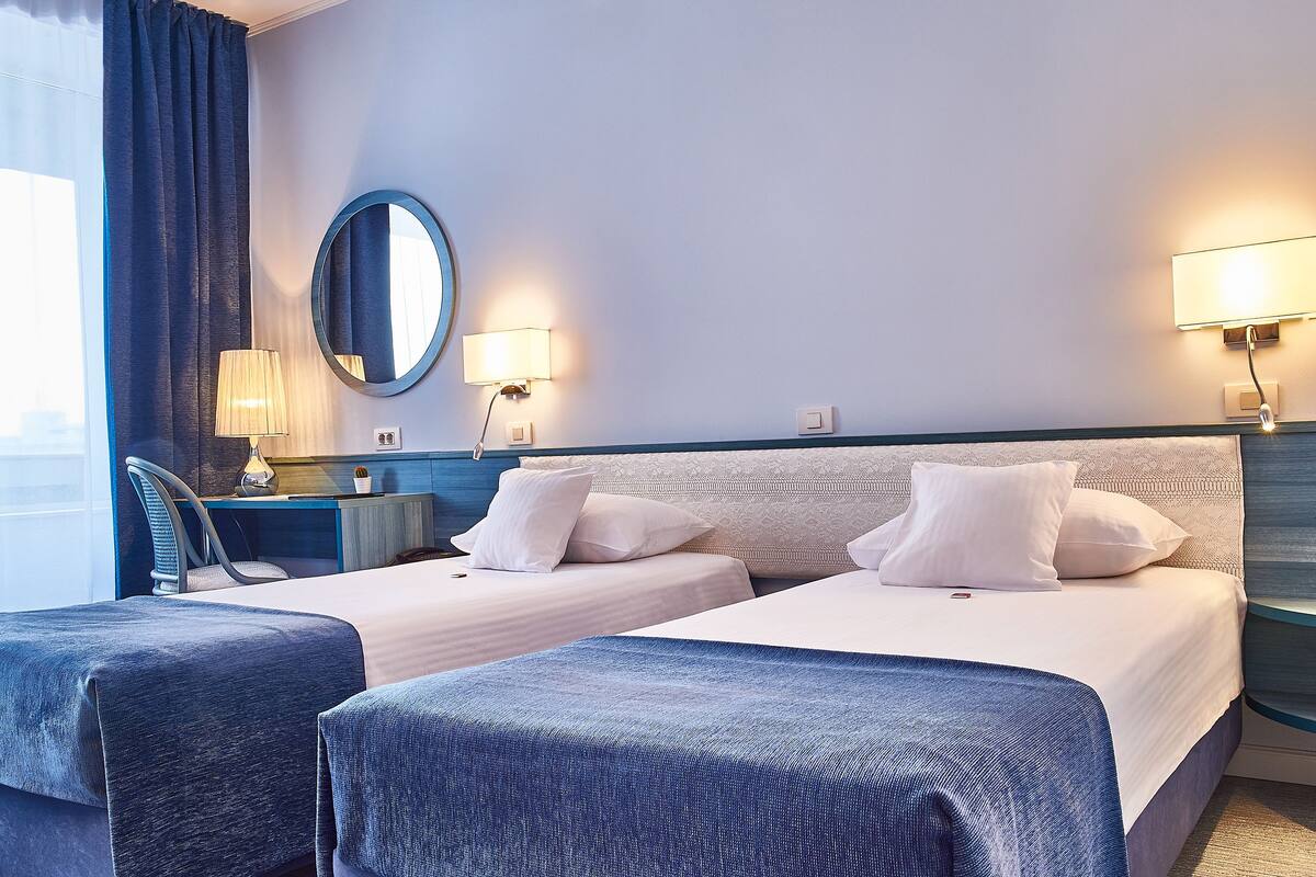 A hotel room with two beds, a desk, a chair, and a round mirror.