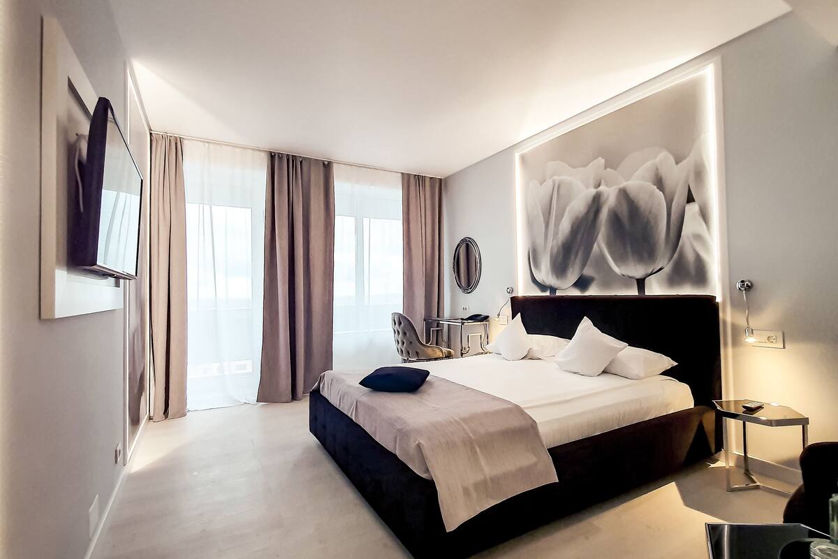 A modern hotel room with a large bed, a flat-screen TV mounted on the wall, a bedside table with a lamp, and a large window with curtains.