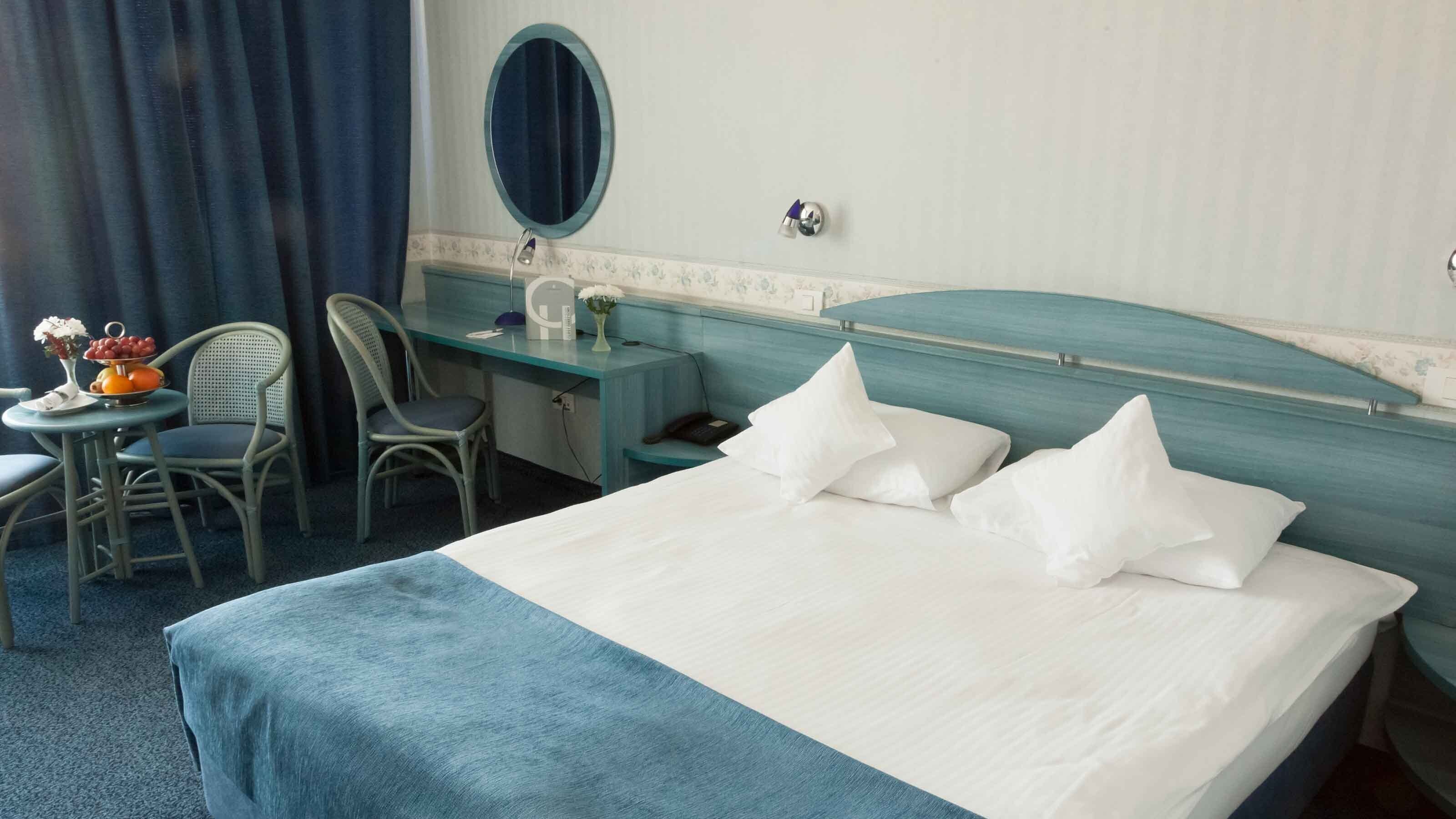 Een hotelkamer met een bed, een bureau, een stoel, een klein tafeltje met fruit en een vaas, en een spiegel aan de muur.