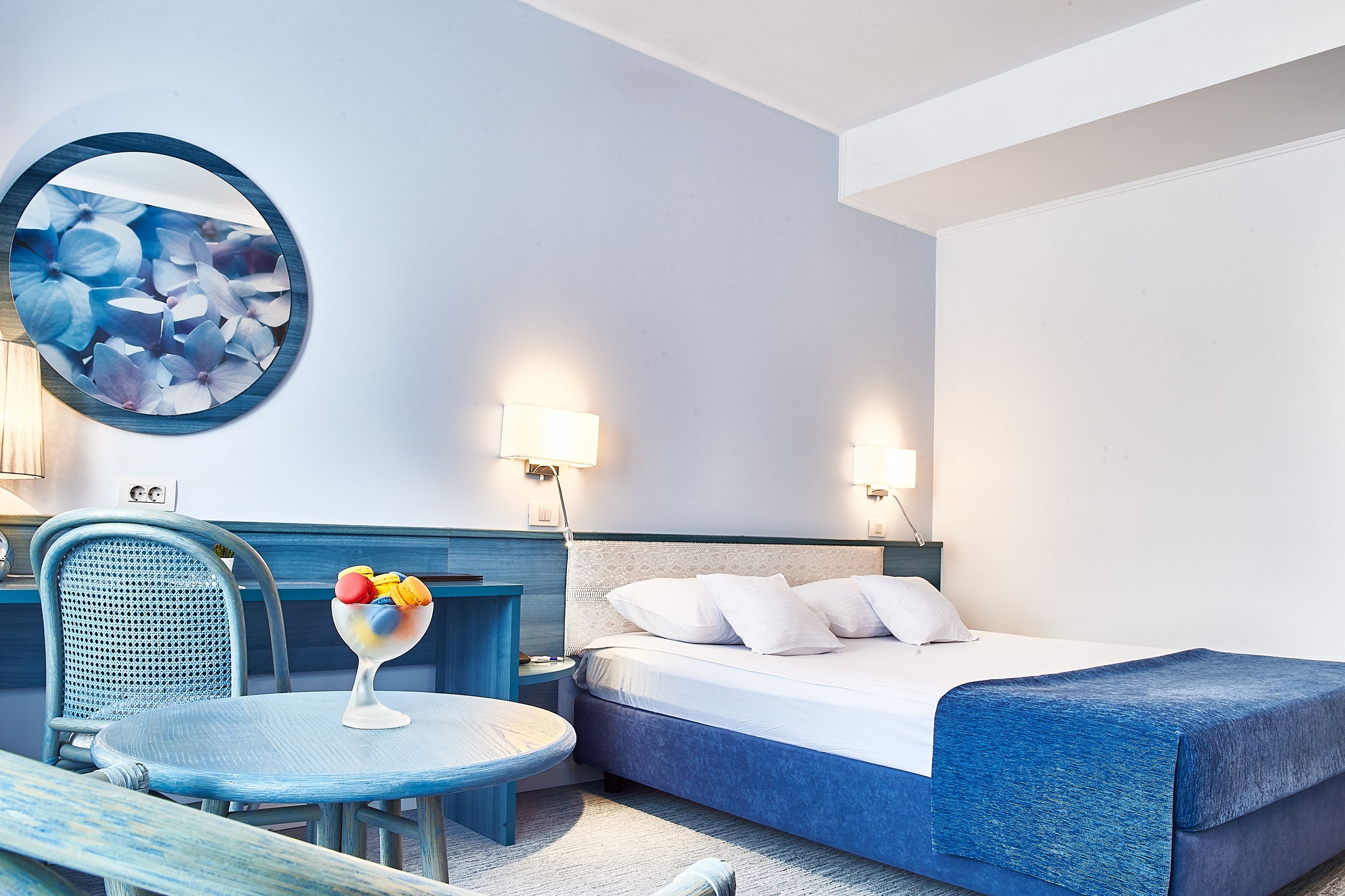 A hotel room with a bed, a round table with a fruit bowl, a chair, and a wall-mounted round mirror.