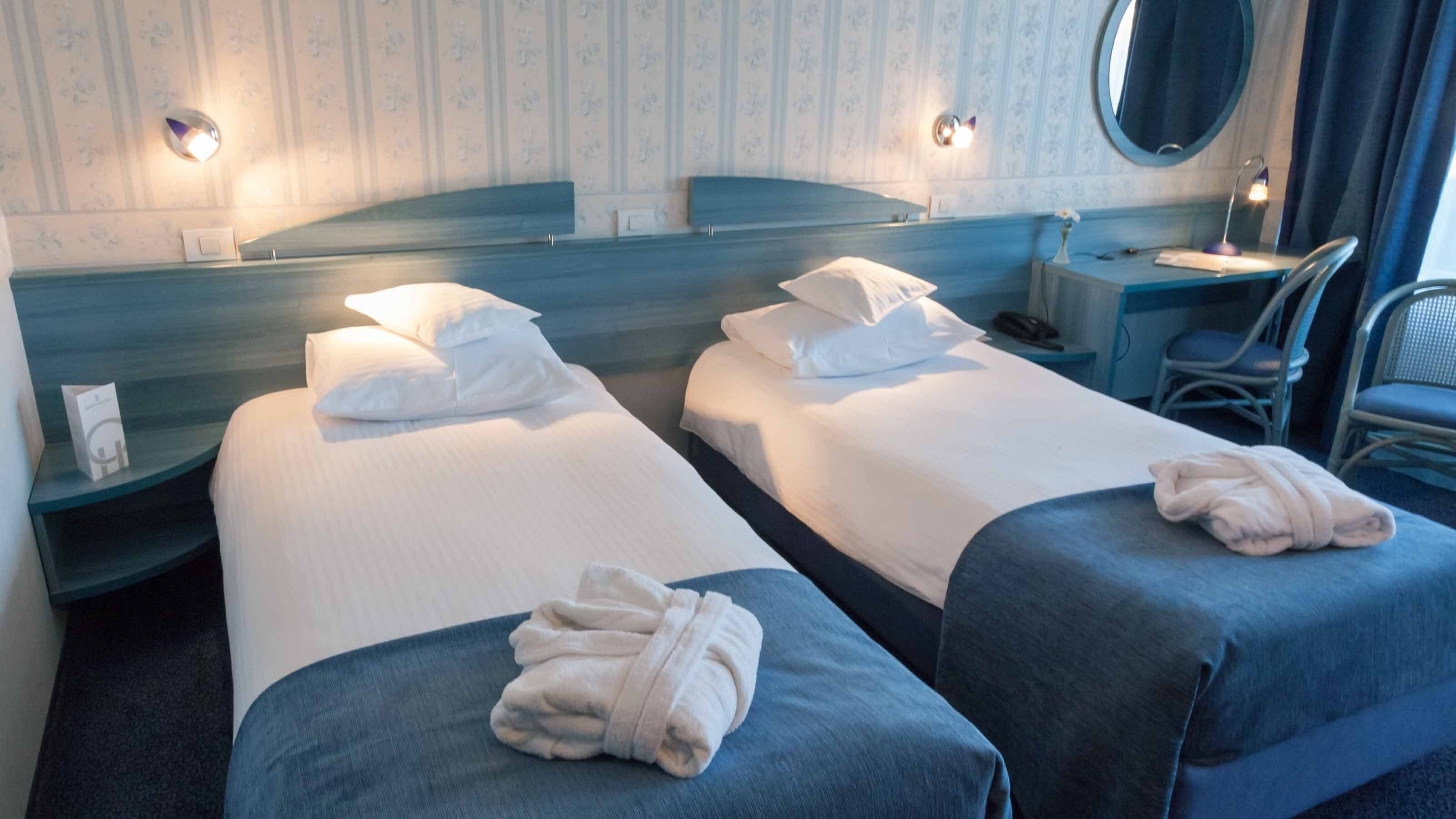 Ein Hotelzimmer mit zwei Betten, jeweils mit weißen Leinen und Kissen, einem runden Spiegel, einem kleinen Tisch mit Lampe und einem Sessel.