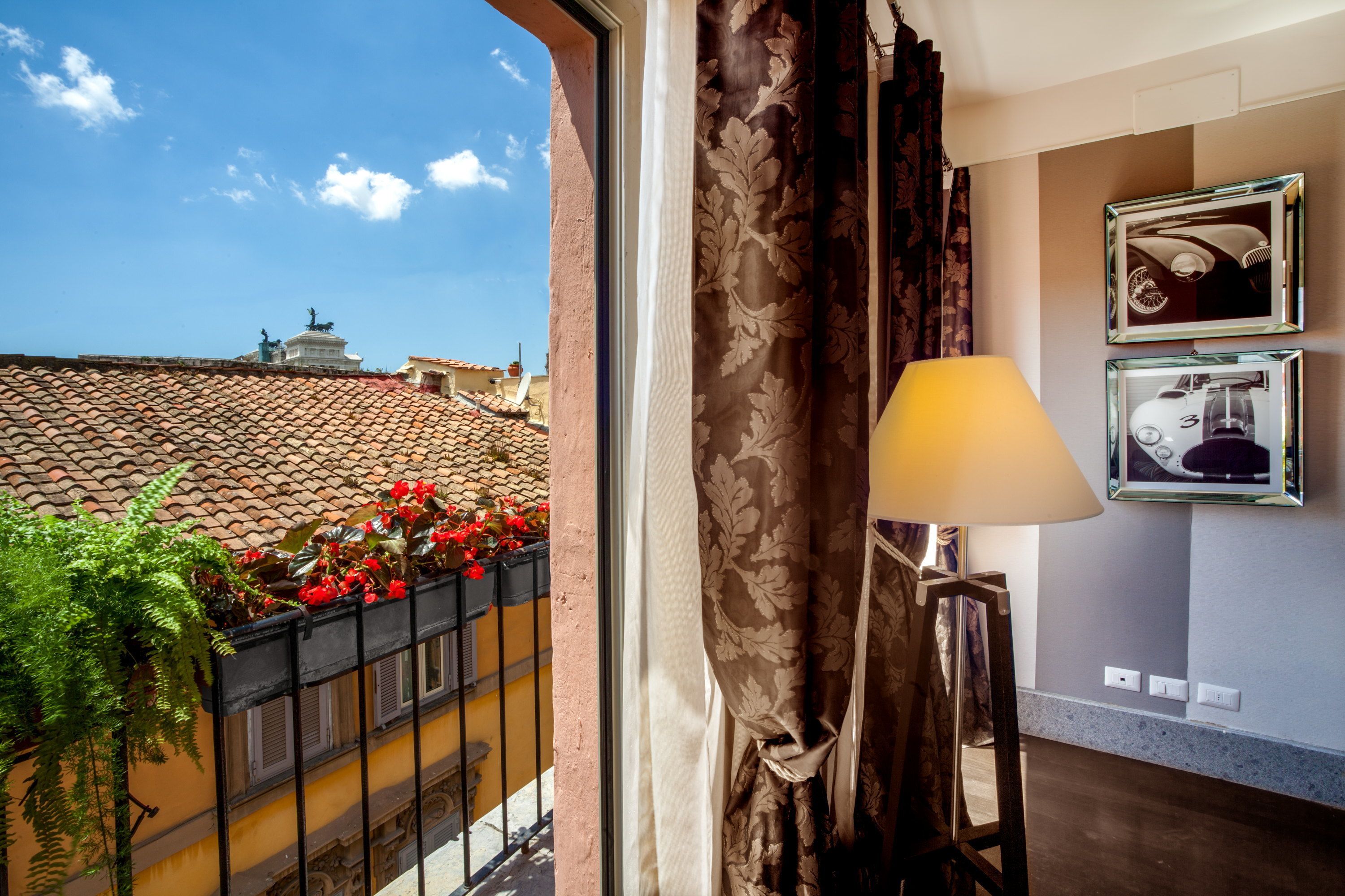 A room with a view of red-tiled roofs and a statue, a lamp, and framed pictures on the wall.