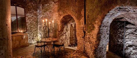 Una habitación con paredes de piedra, una mesa, sillas, un candelabro y un ventanal.