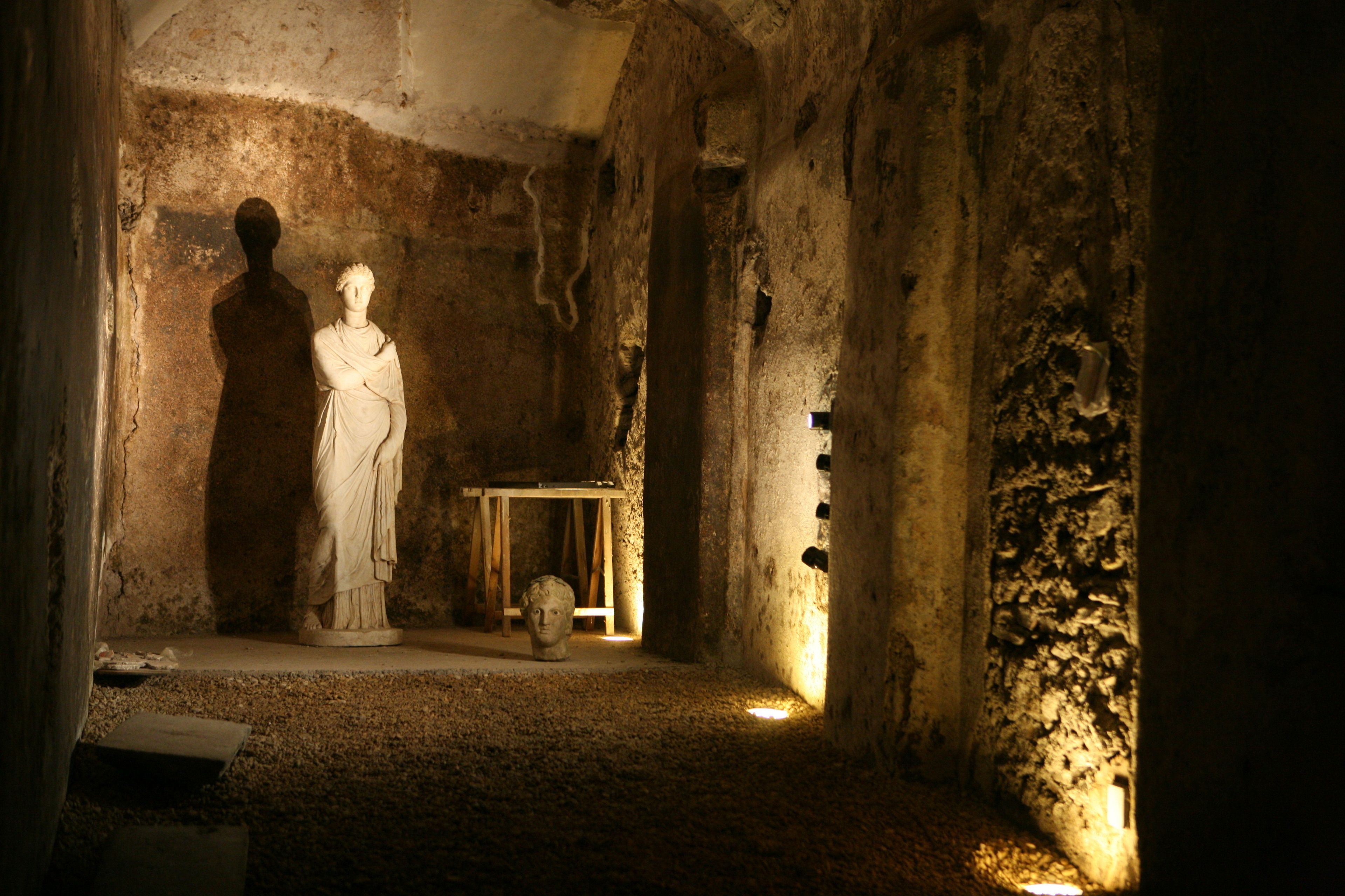 A dimly lit interior with a statue and a bust on pedestals, stone walls, and a small table with a lamp.