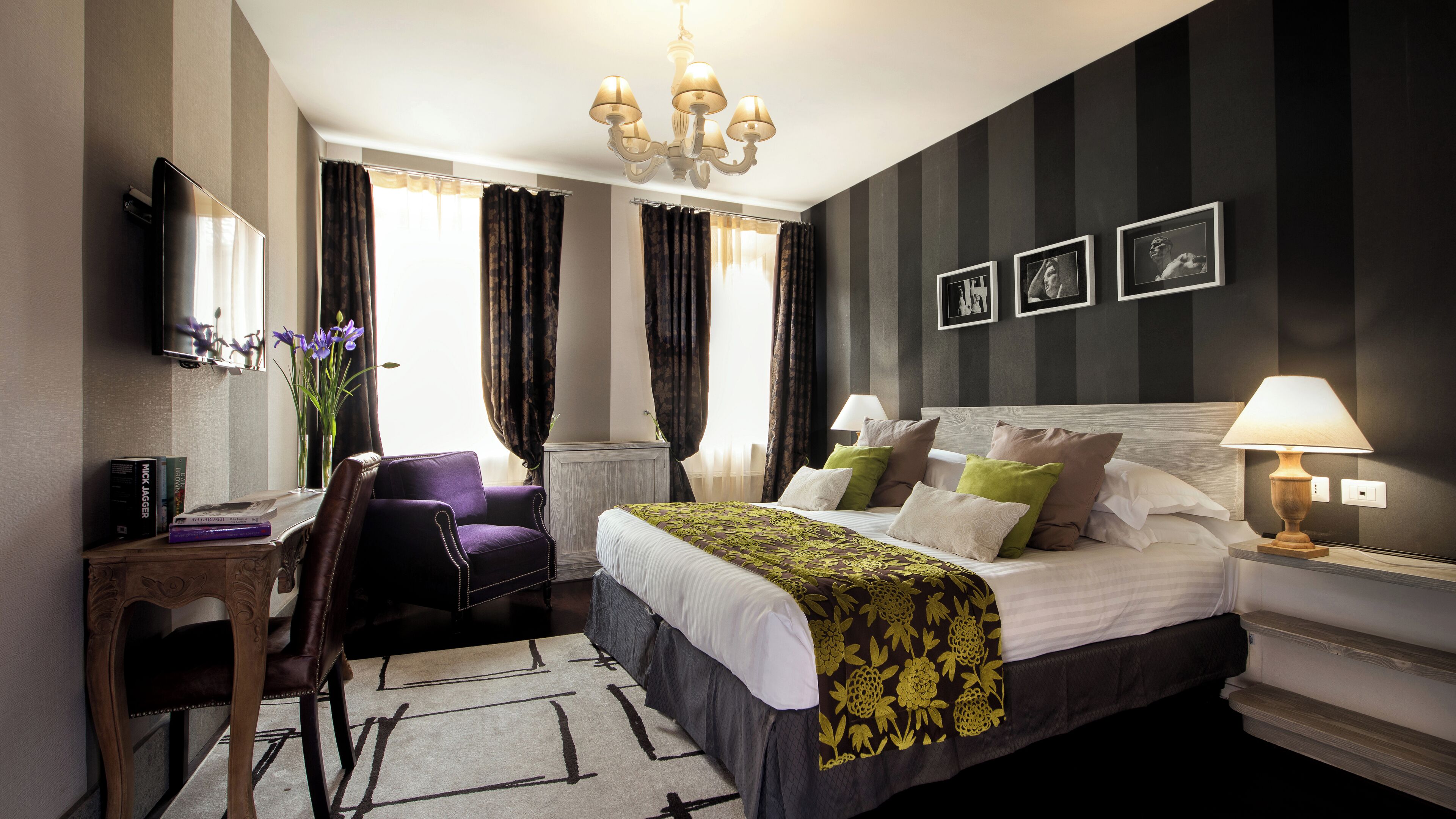 A hotel room with a bed, a desk, a chair, a chandelier, and a wall with framed pictures.