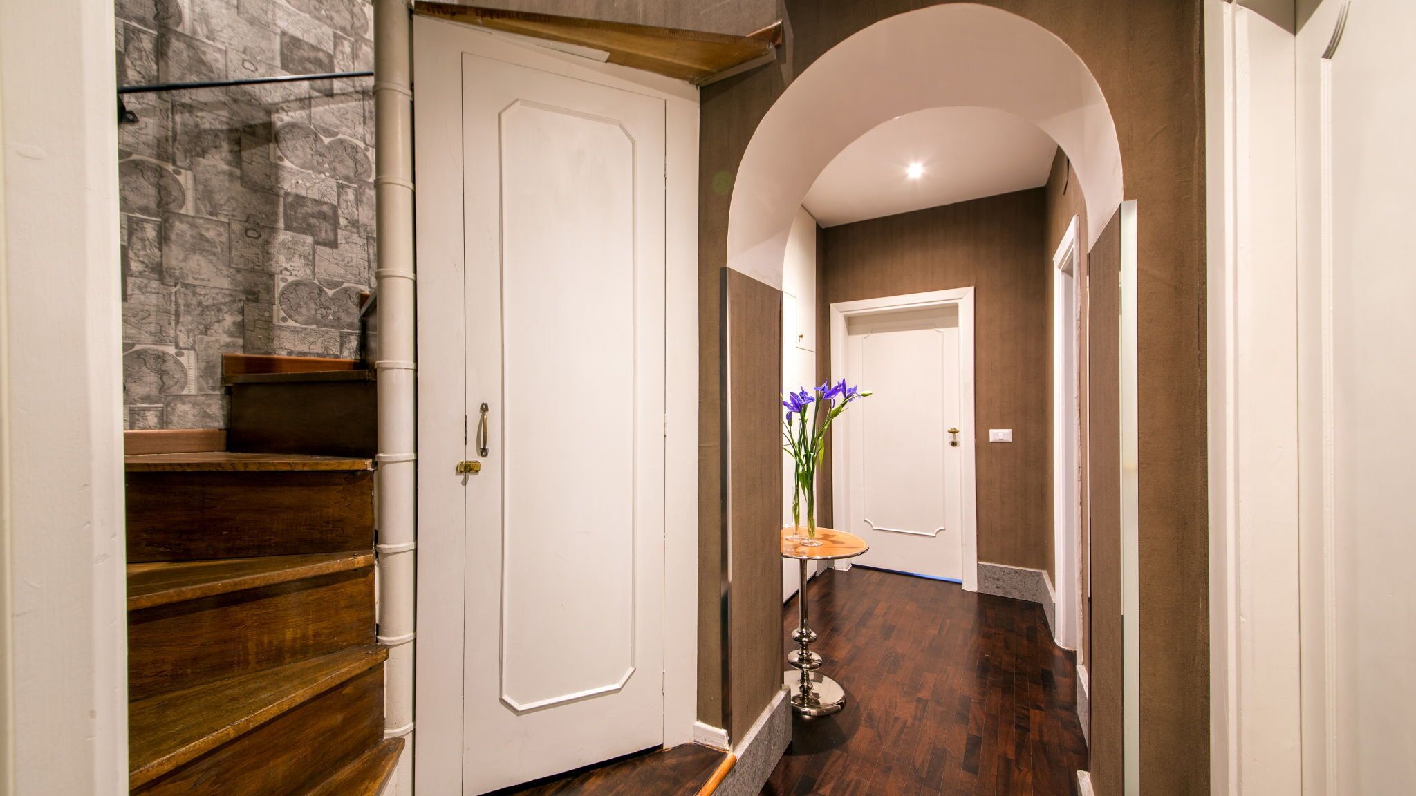 Un couloir avec un mur en pierre, des marches en bois, une porte blanche et un vase rempli de fleurs.