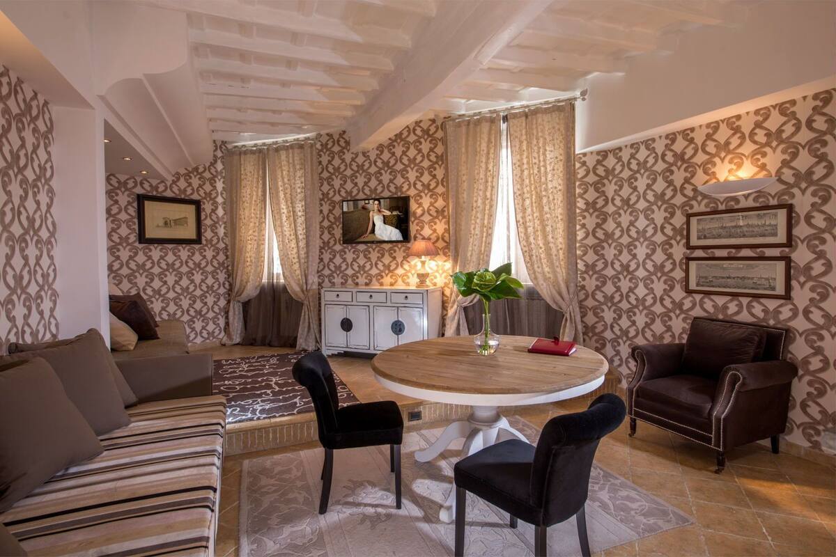 Un salon avec une table ronde en bois, deux chaises noires, un canapé, une table basse et un téléviseur fixé au mur.