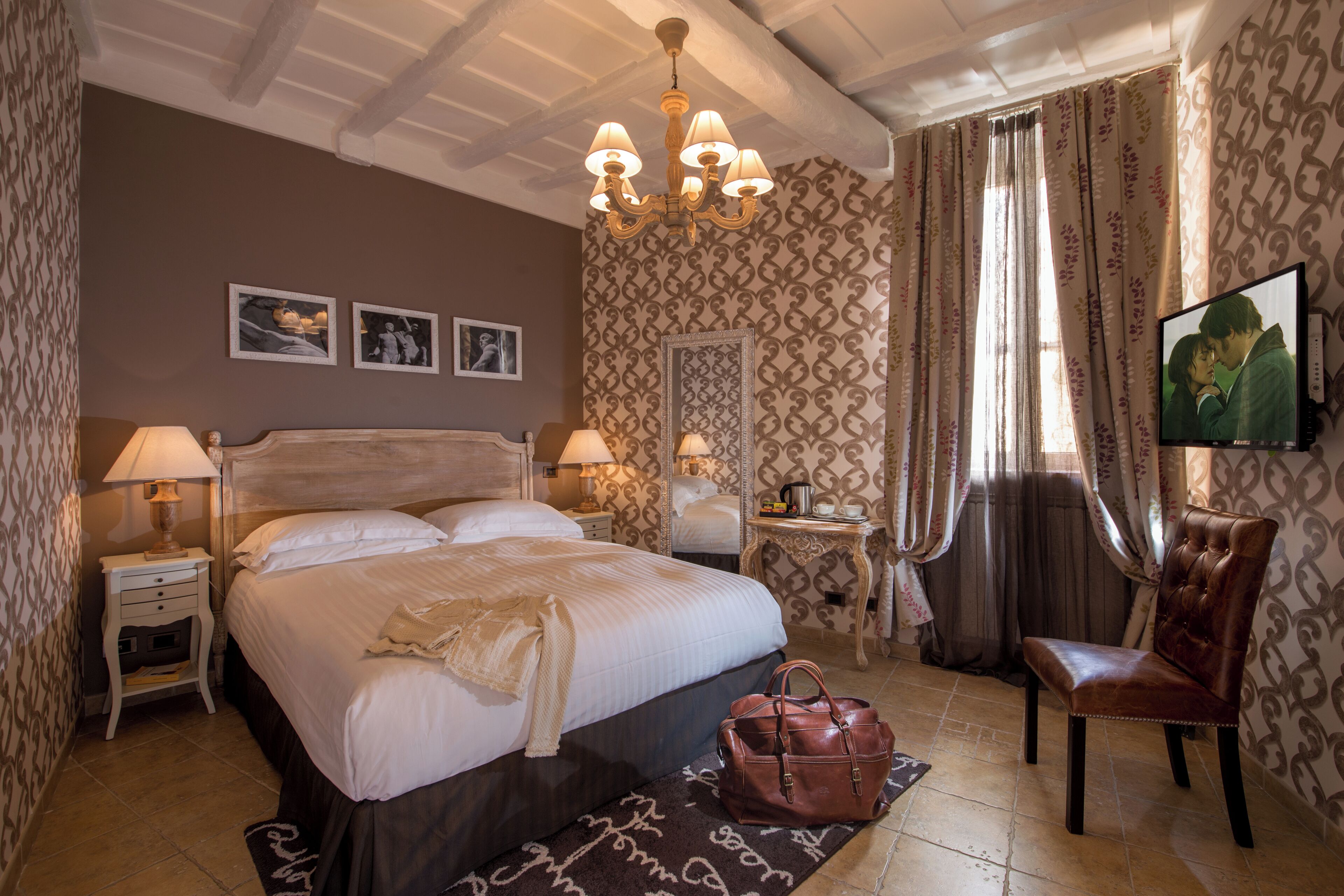 Une chambre d’hôtel avec un grand lit, deux tables de chevet, un lustre, une télévision et un papier peint à motifs.