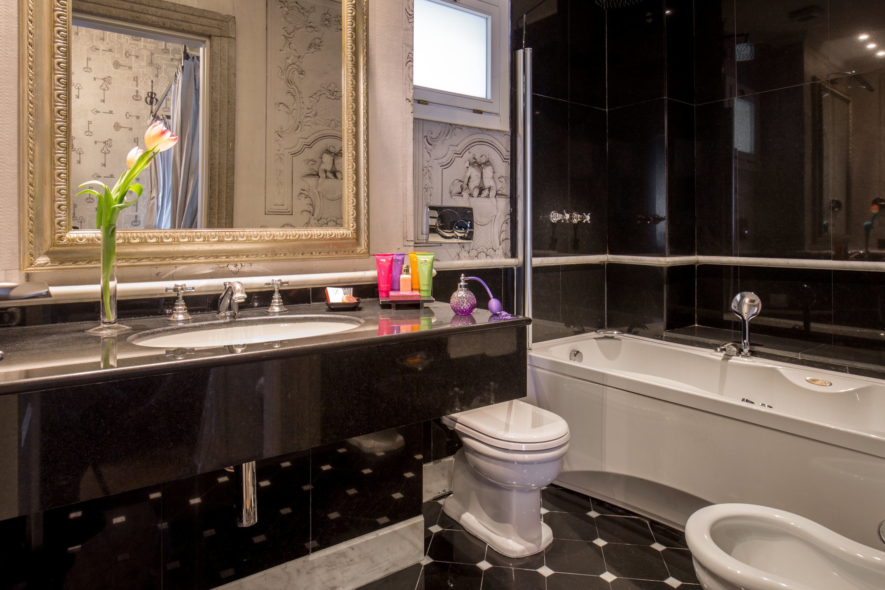 A modern bathroom with a black and white tiled floor, a white toilet, a white bathtub, and a large mirror with a gold frame.