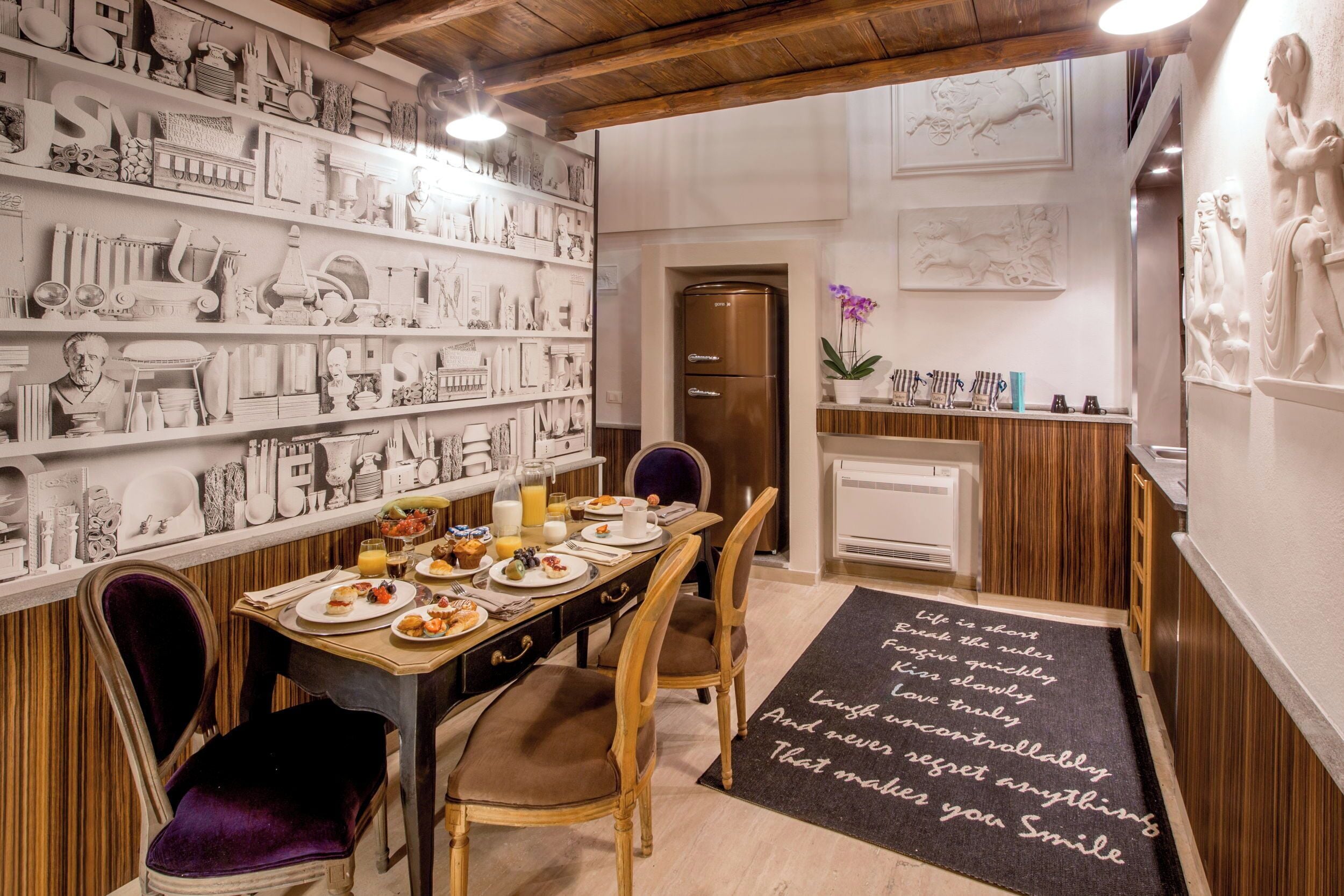 On aperçoit un coin repas avec une longue table dressée pour quatre personnes, entourée de chaises. Un réfrigérateur est visible à l’arrière-plan, et les murs sont décorés de diverses sculptures et œuvres d’art blanches.