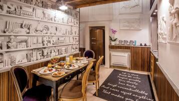 On aperçoit un coin repas avec une longue table dressée pour quatre personnes, entourée de chaises. Un réfrigérateur est visible à l’arrière-plan, et les murs sont décorés de diverses sculptures et œuvres d’art blanches.