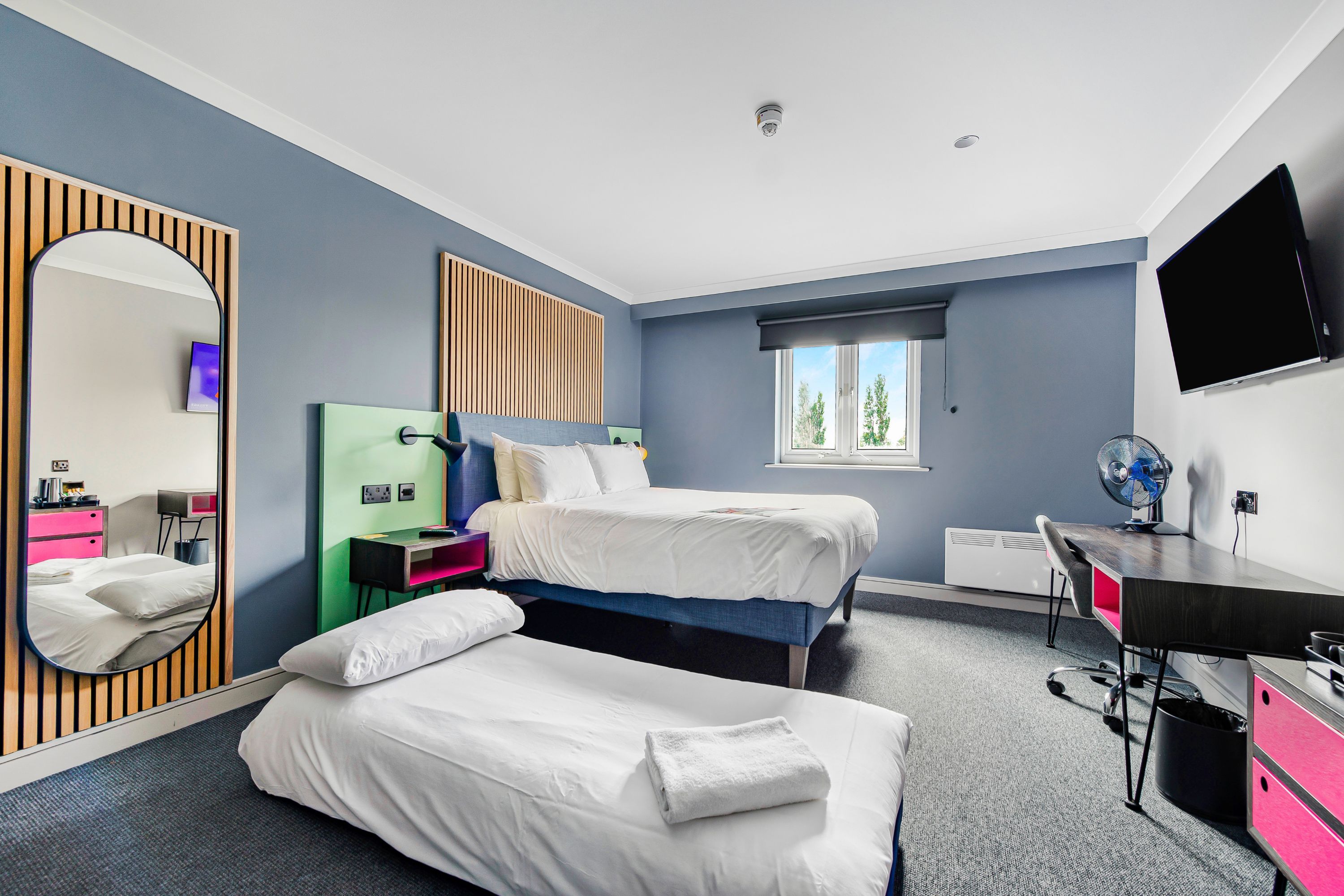 A modern hotel room with two beds, a desk, a mirror, and a TV.