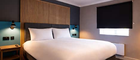 A hotel room with a large bed, wooden headboard, two bedside tables with lamps, a window with a blind, and a wall-mounted thermostat.