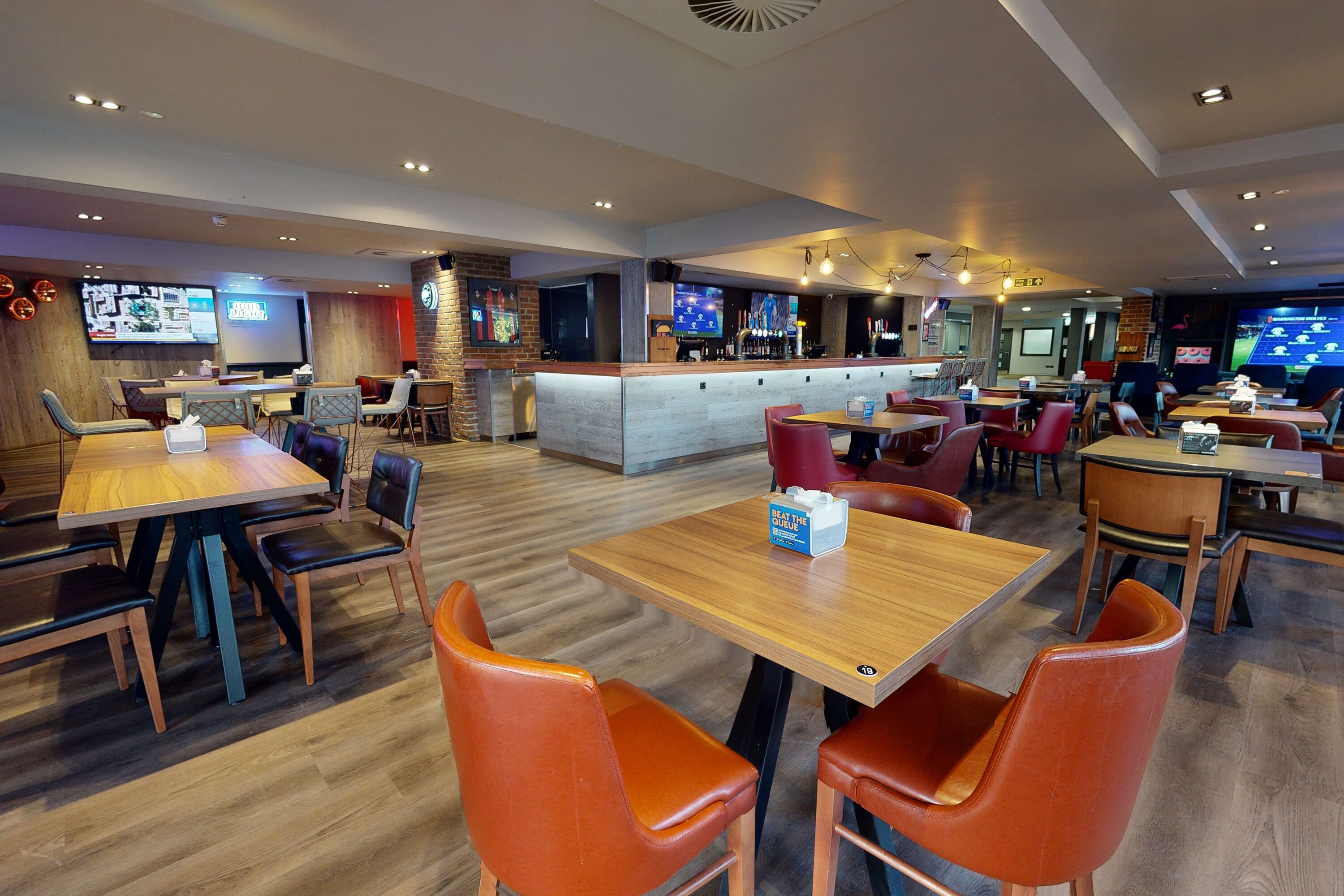 A restaurant with wooden tables and orange chairs, a bar area, and multiple screens displaying content.