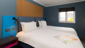 A hotel room with a large bed, a wooden headboard, a bedside table, a nightstand, a window with a view, and a wall-mounted light fixture.